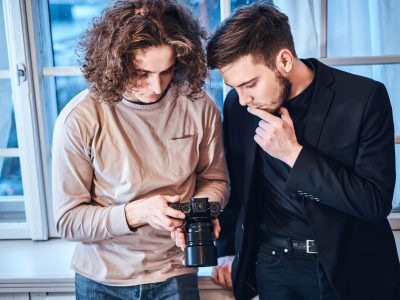 Close up photo of professional curly photographer man showing photos on digital camera to suit dressed custumer at photo studio indoors